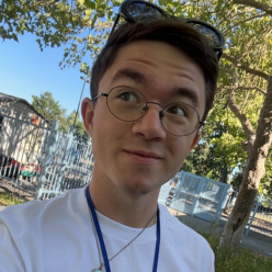 White male in a white t shirt shirt with brown hair and round glasses. Standing in front of a blue fence and a tree. 